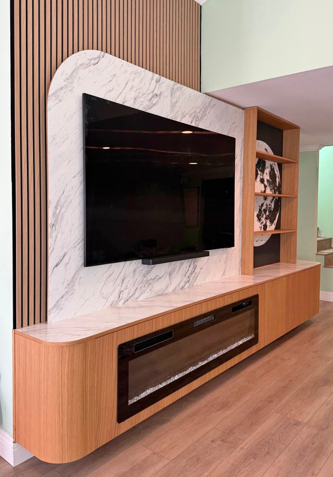 Custom media wall with wood slat accent, marble-look TV panel, electric fireplace, built-in shelves, and wood cabinets in Houston home